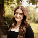 A photo of a woman with brown hair with a tree in the background.